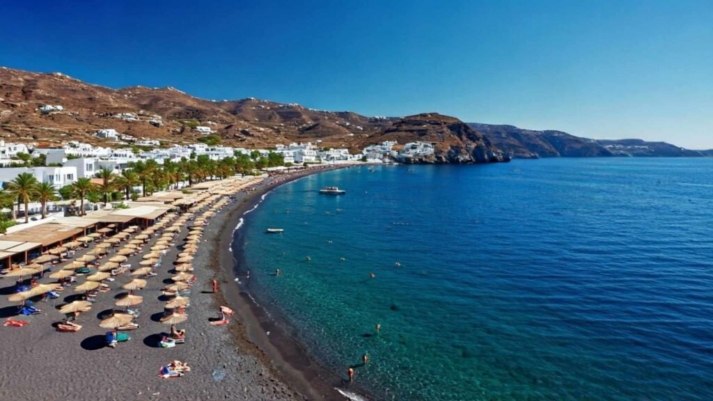 Aerial view of a black sand beach with sunbeds and umbrellas along the clear Aegean Sea in Santorini