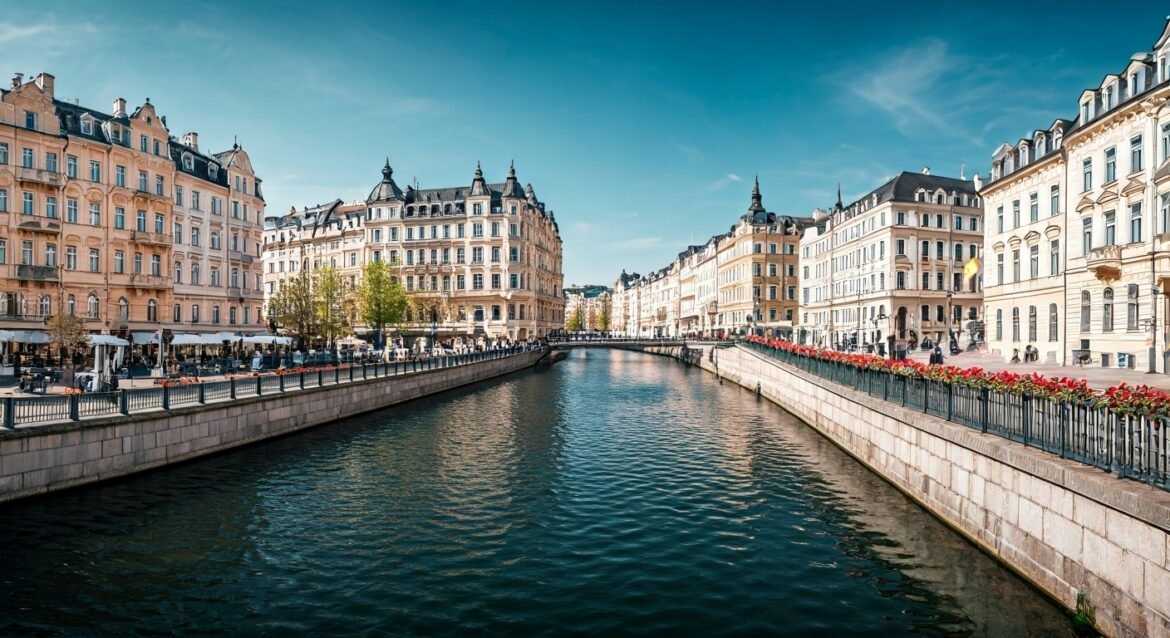 A high-resolution daytime view of Karlovy Vary, Czech Republic, featuring elegant neo-Baroque architecture along a canal lined with cafés and flower-filled railings.