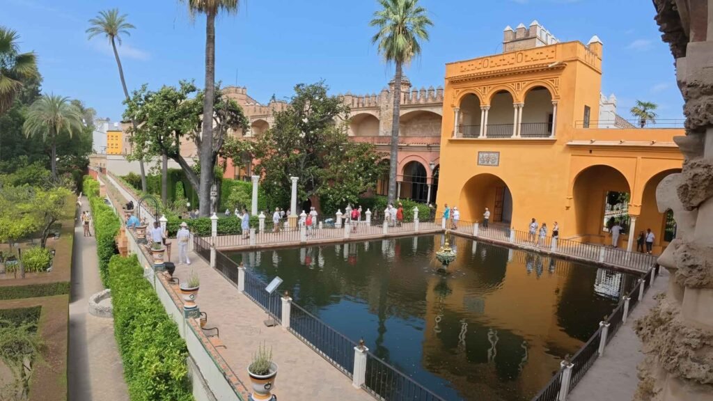 Wide-angle view of Real Alcázar Palace courtyard in Seville, showing Moorish arches, tilework, and reflection pool under golden sunlight