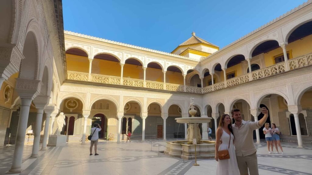 A young couple takes a selfie near the arches of Casa de Pilatos in Seville, Spain, with the historic courtyard and central fountain visible in the background.