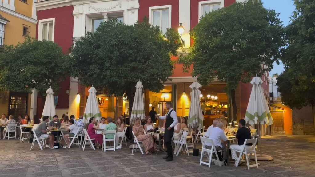 Evening outdoor restaurant scene in Santa Cruz, Seville, with people dining under orange trees and warm golden light on cobblestone streets.