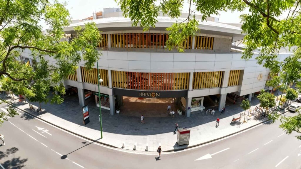A sunny street view of Nervión Plaza Shopping Center in Seville with people walking by and the modern white-and-gold facade captured from a slightly elevated angle.