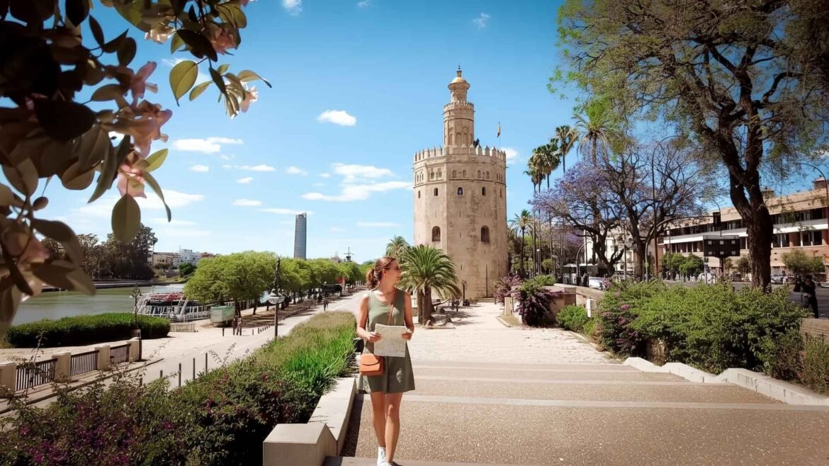 A young woman wearing a straw hat walks along the riverside promenade near Torre del Oro in Seville, Spain, on a sunny day.