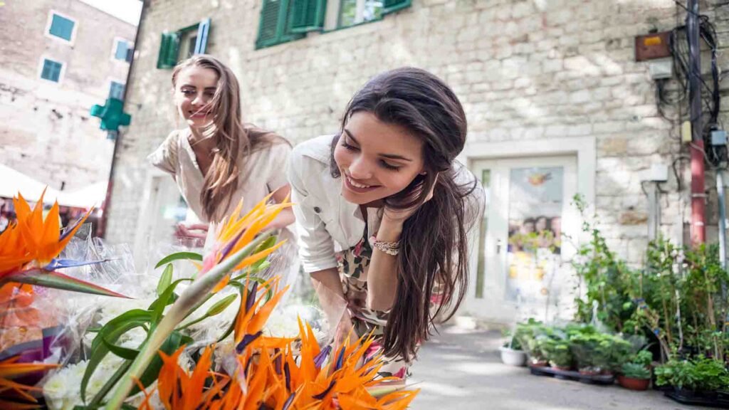 Two young women tourists admiring colorful flowers at Campo de’ Fiori market in Rome on a lively day