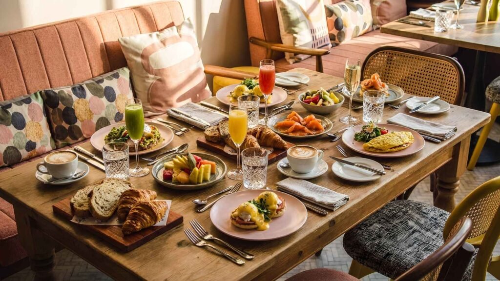 A beautifully arranged luxury hotel breakfast table in Bangkok featuring eggs benedict, omelette, croissants, fresh bread, fruit plates, and colorful juices in a sunlit dining room.