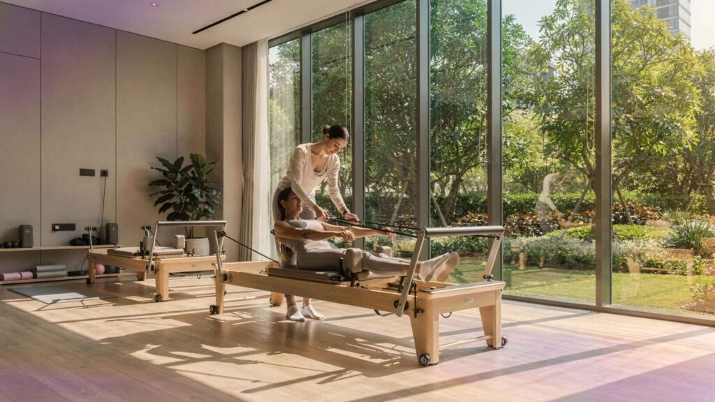 A pilates instructor assisting a resident during a reformer pilates session inside a modern residential wellness studio with garden views.