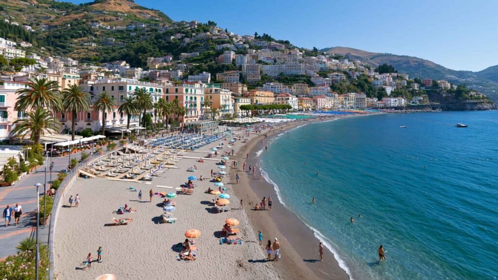 Wide view of Maiori Beach with the seafront promenade, palm trees, clear water, and the hillside town on the Amalfi Coast.