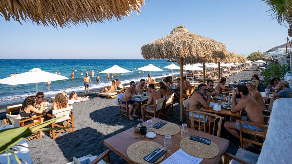 Beachfront restaurants near Santorini beaches with outdoor dining, black sand shoreline, and busy seaside atmosphere