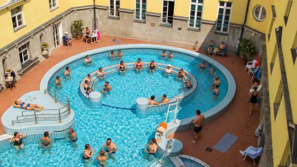 High-angle view of visitors relaxing and swimming in a circular outdoor thermal pool enclosed by historic yellow building walls.