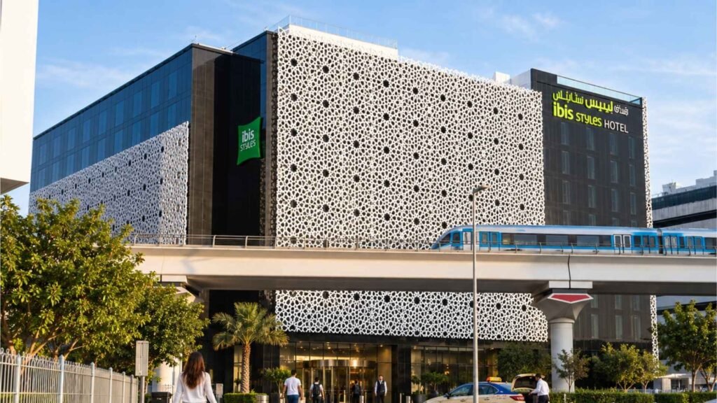 Exterior view of ibis Styles Dubai Airport with its distinctive white geometric façade, an elevated Dubai Metro train, arriving travellers, an airport taxi and passing traffic near the hotel entrance.