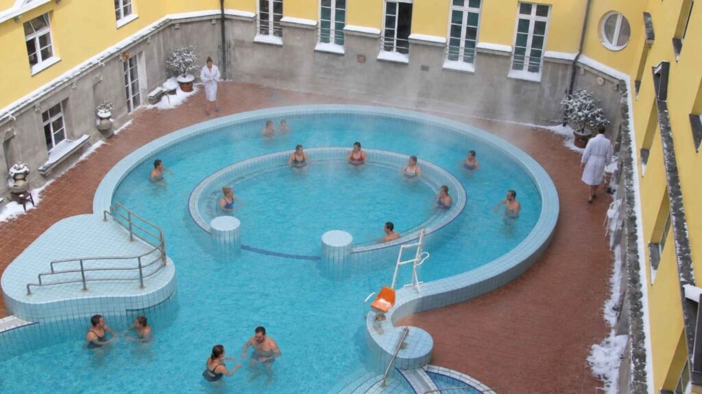 Bathers in Lukács Thermal Bath’s outdoor pool, with light steam and snow along the surrounding yellow courtyard walls.