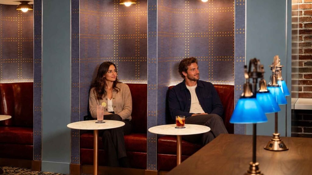 Two travellers seated in separate leather lounge booths with marble tables, colourful drinks and blue lamps in the foreground.