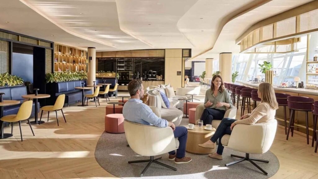 Three friends chatting in cream armchairs around a wooden coffee table in a bright lounge with large windows and burgundy bar stools.