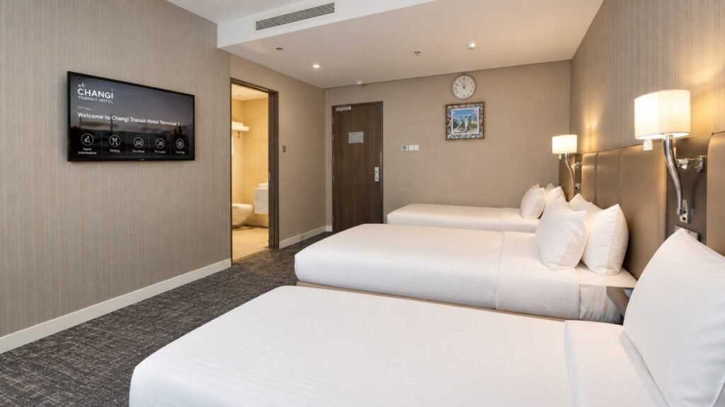 Family Room at Changi Transit Hotel T3 with three beds, white bedding, bedside lamps and an open bathroom doorway.