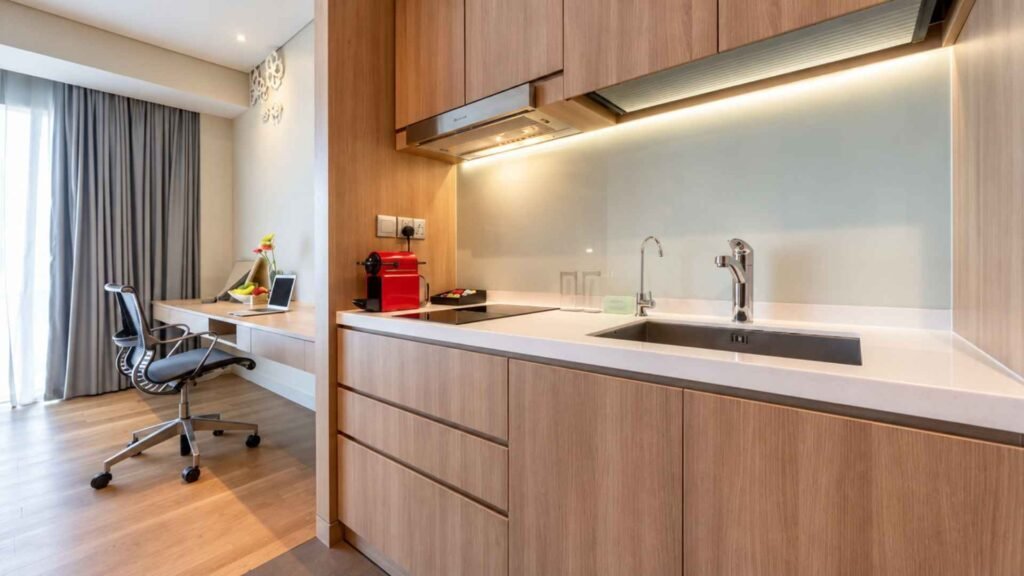 Kitchenette with wooden cabinets, a sink and a red coffee machine beside a window-side work desk at Dorsett Changi City Singapore.