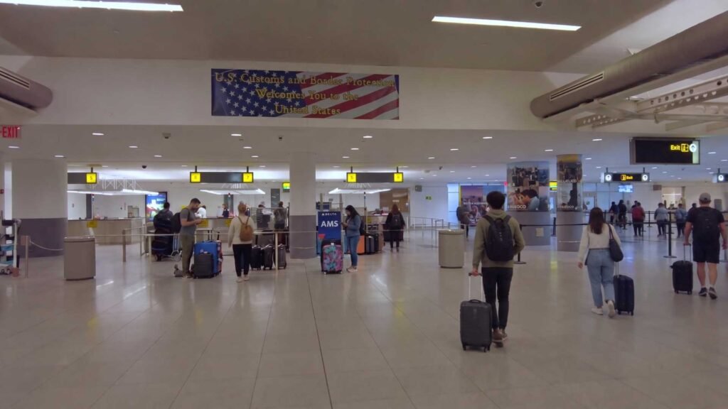 Travellers with suitcases walking towards exit signs beneath a U.S. Customs and Border Protection welcome banner at JFK.