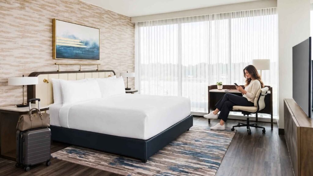 Hyatt Regency JFK hotel room with a woman checking her phone by the window and luggage beside the bed.