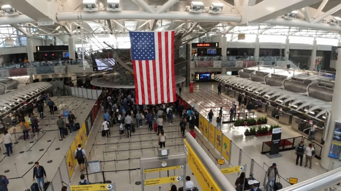 JFK Airport terminal interior with a large American flag and passengers queuing beneath the steel-beamed ceiling.