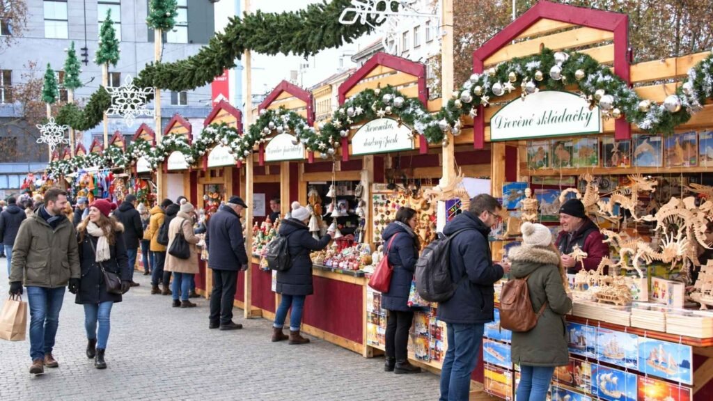 Visitors browsing wooden toys and Christmas gifts at decorated market stalls in Prague during the day.