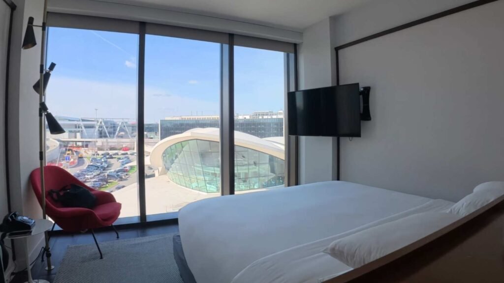TWA Hotel room with a bed, red armchair and floor-to-ceiling windows overlooking the TWA Flight Center.
