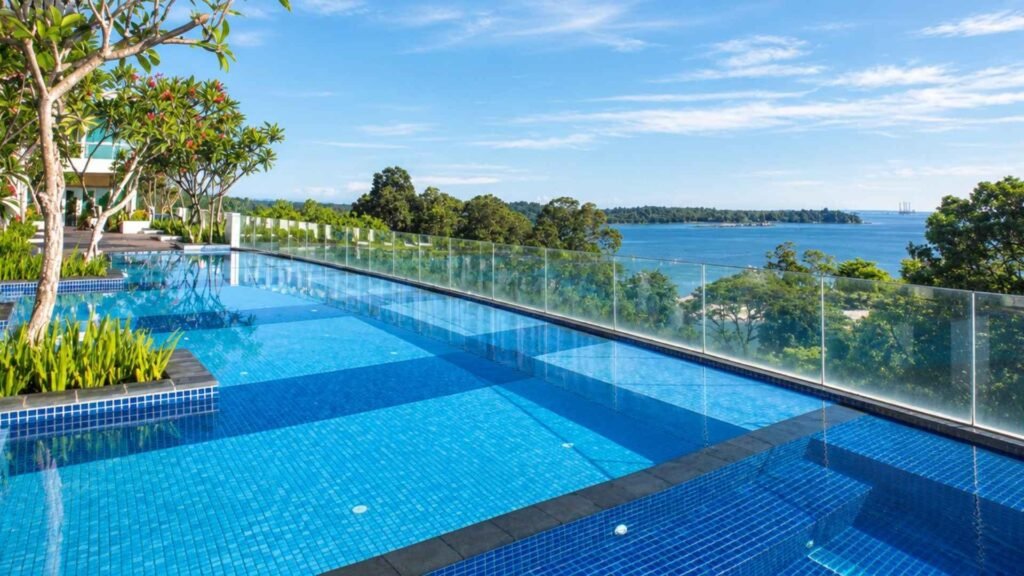 Blue-tiled swimming pool at Village Hotel Changi, with glass railings overlooking green treetops and the sea.