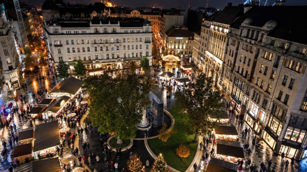 Elevated view of Vörösmarty Square Christmas market in Budapest, with illuminated wooden stalls, visitors and lights reflected on wet paving.